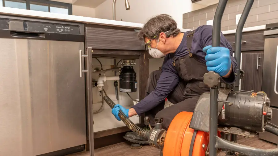 A masked plumber feeding a drain-cleaning auger cable beneath a kitchen sink toward the garbage disposal